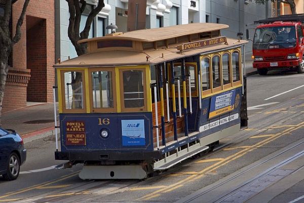 A yellow and blue cable car numbered 16 travels on a street with tracks. A red vehicle is visible in the background.