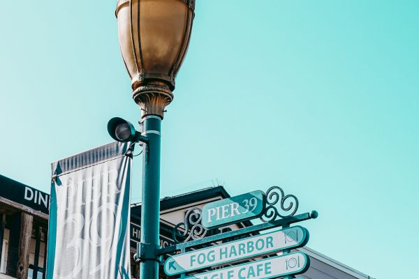 The image features a street lamp post with directional signs in front of shops, set against a clear blue sky.