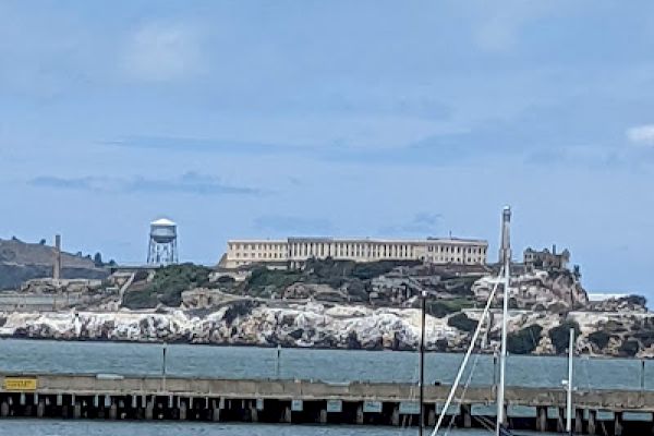 This image shows a coastal scene with a historic building in the background, a pier, and two boats in the water under a blue sky with light clouds.