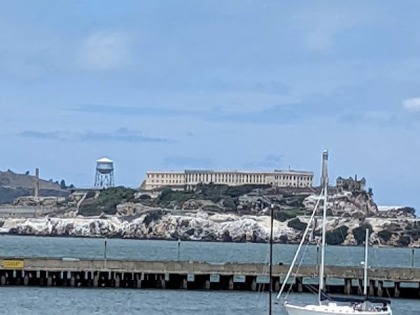 This image shows a coastal scene with a historic building in the background, a pier, and two boats in the water under a blue sky with light clouds.