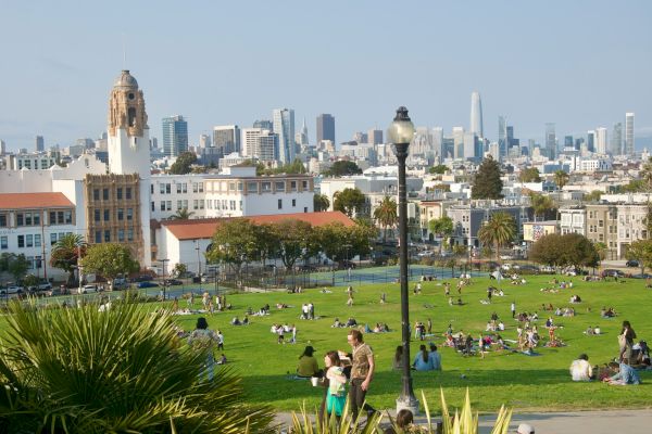 People relaxing on a green lawn with a city skyline in the background, including historic buildings and skyscrapers under a clear sky.