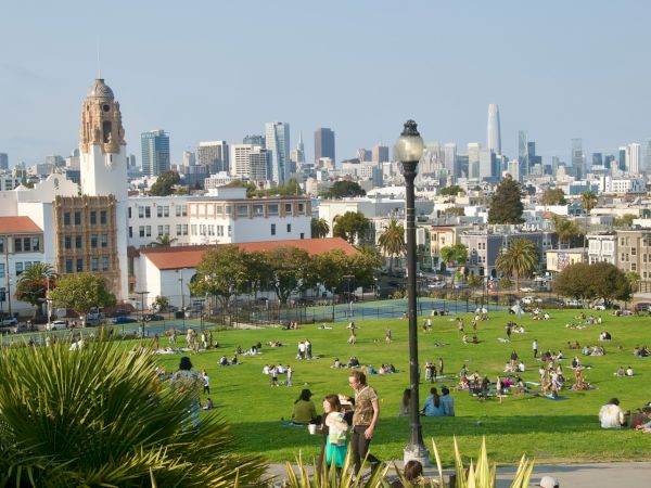 People relaxing on a green lawn with a city skyline in the background, including historic buildings and skyscrapers under a clear sky.