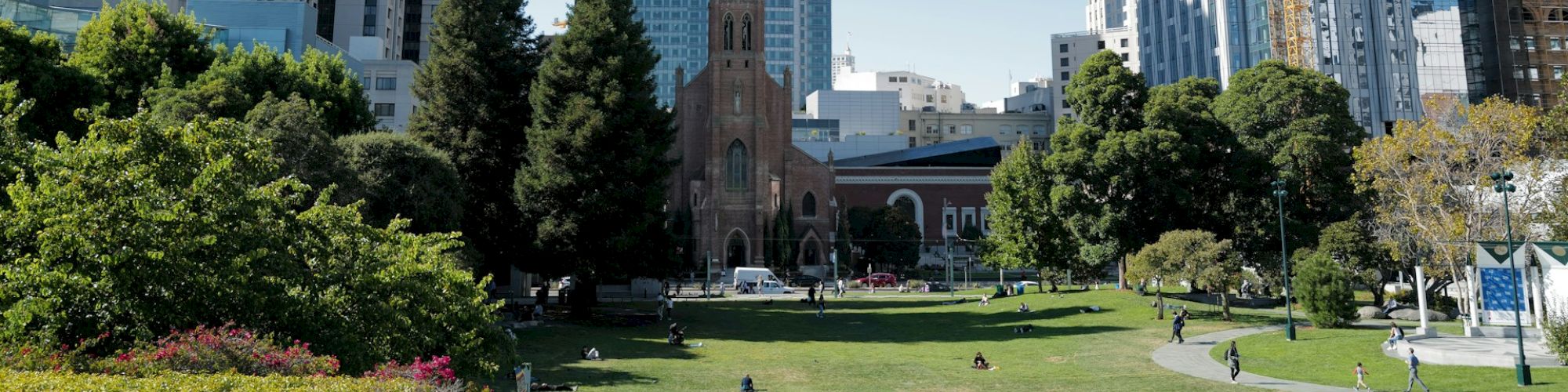 A cityscape featuring modern high-rise buildings, a historic church, and a park with greenery and people enjoying the outdoors.