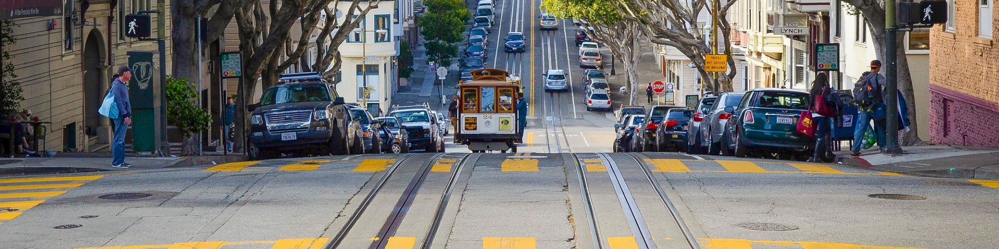 A street in a city with yellow pedestrian lines, cable car tracks, parked cars, and trees lining both sides of the road with a cable car in the distance.