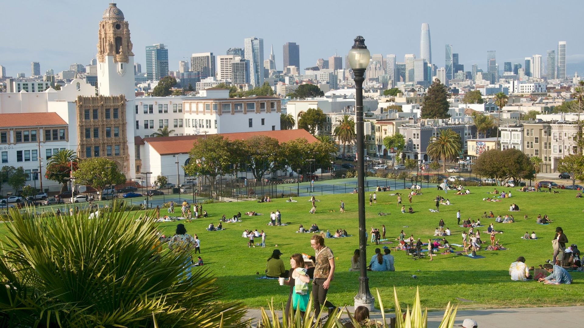 People relaxing in a green park with a cityscape in the background, featuring buildings and skyscrapers, under a clear sky with a lamppost in view.