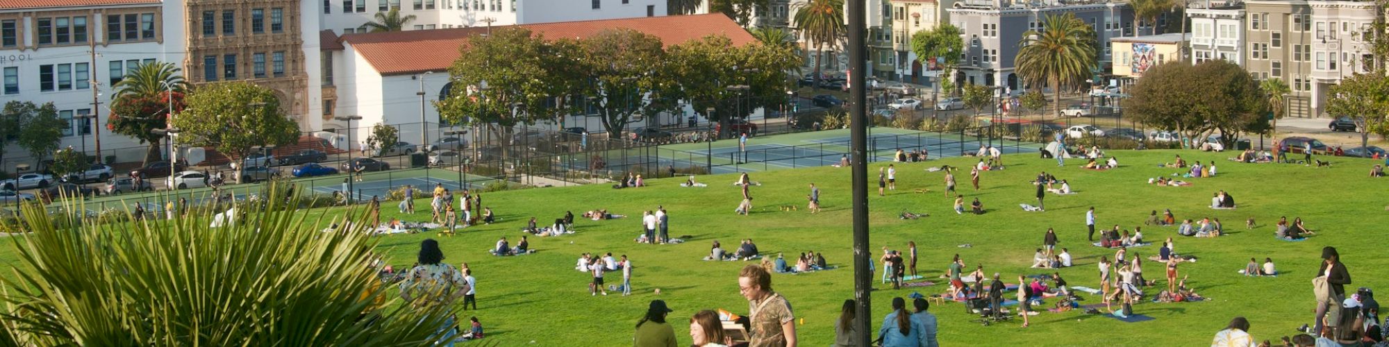 People relaxing in a green park with a cityscape in the background, featuring buildings and skyscrapers, under a clear sky with a lamppost in view.