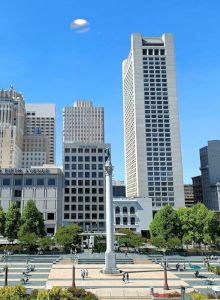 The image shows a cityscape with various tall buildings, a central column monument, green trees, and an outdoor urban area under a clear blue sky.