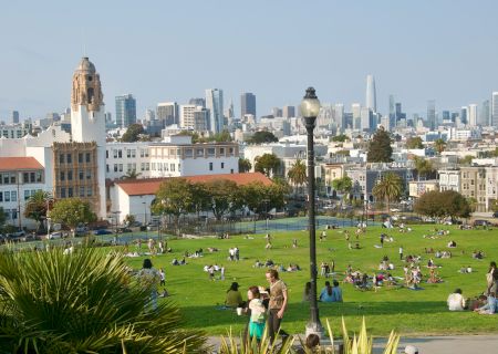 A park filled with people relaxing on a sunny day, city skyline and historic buildings in the background, palm trees in the foreground.