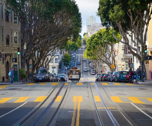 A streetcar travels up a hilly, tree-lined street in a city, with cars parked on both sides and buildings lining the road.