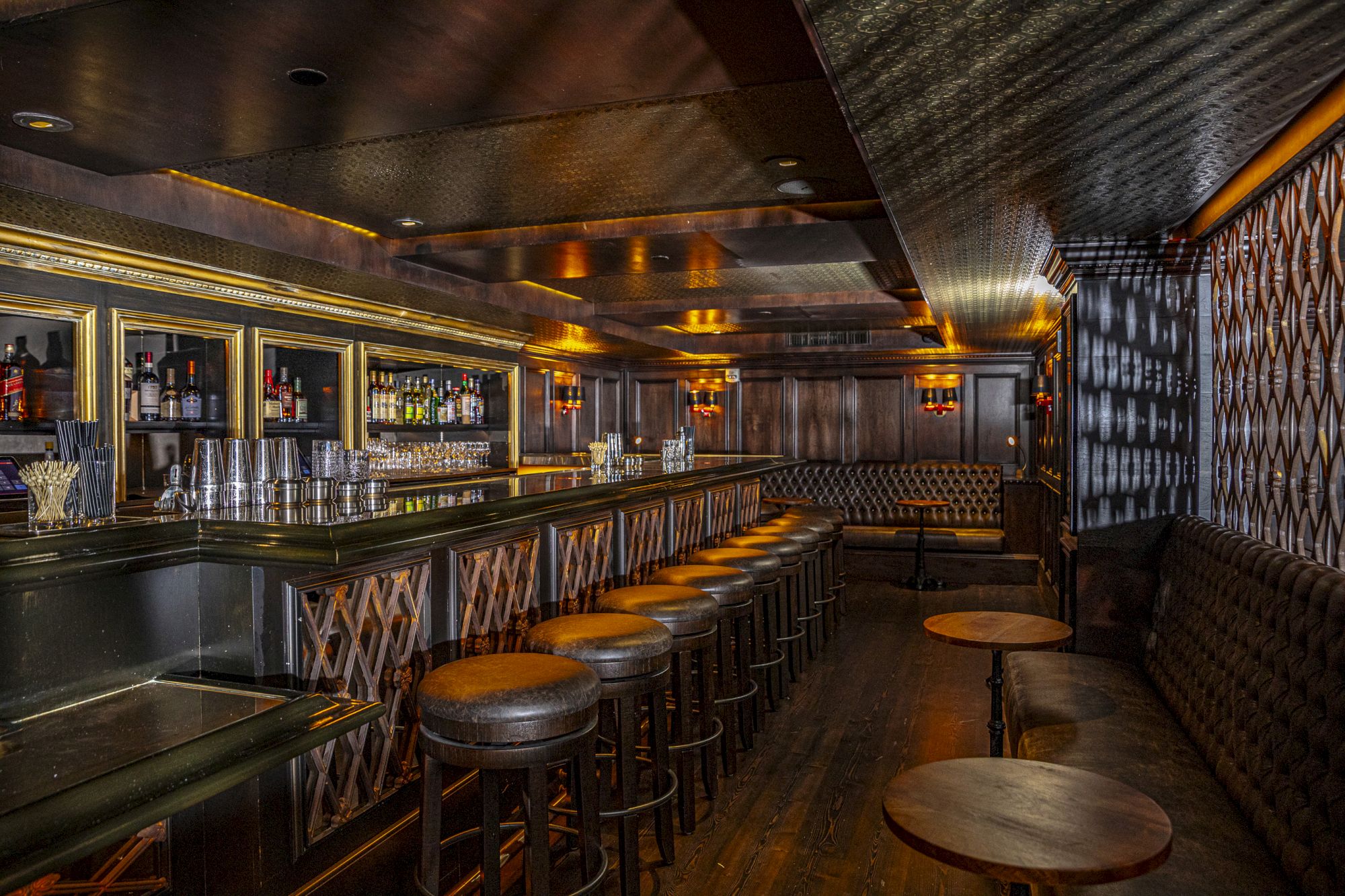 The image shows an empty, dimly-lit bar with a long counter, bar stools, and tables along one side. The decor features dark tones and ambient lighting.