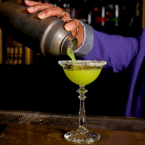 A bartender pours green cocktail from a metal shaker into a stemmed glass, with a blurred bar in the background.