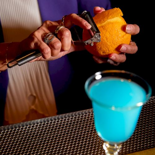 A bartender&rsquo;s hands slice or peel an orange with a small tool over a bar while a blue drink sits nearby.