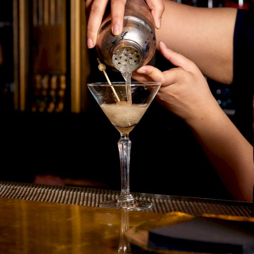 A bartender pours a drink from a metal shaker into a martini glass at a bar.
