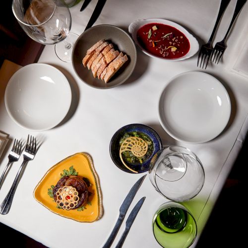 A plated dinner on a white table: bread slices, red soup, greens, sauces, and glasses of wine.