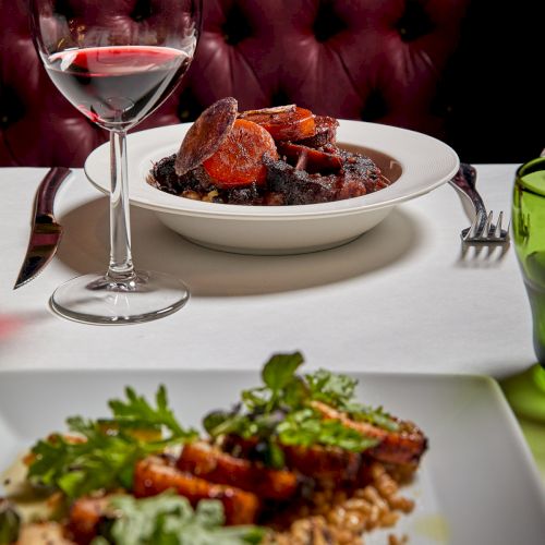 A dining table scene with a glass of red wine and a bowl of roasted meat/sausages, set against a red leather backdrop.