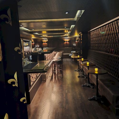A dimly lit bar lounge with leather booths, small round tables, bar stools, and warm ceiling lights.