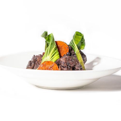 A white plate with a gourmet dish of braised meat, roasted carrots, and leafy greens arranged artistically.