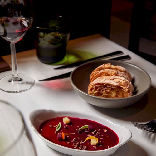 A table set with red soup in a bowl, a crusty bread roll, and a glass of red wine.