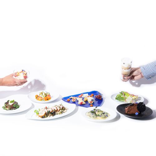 The image shows a variety of dishes including desserts, salads, and entrees. Two hands are reaching in from either side holding desserts.