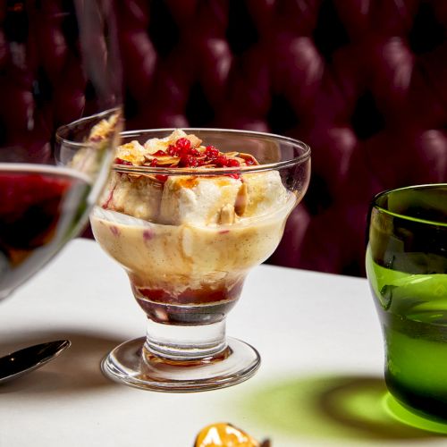 A glass parfait dessert with layered cream, fruit, and nuts sits on a table beside a green drink, with a red-lit backdrop behind.