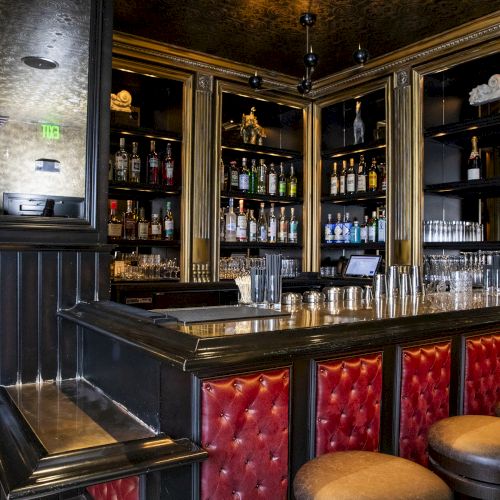 A stylish bar with red-padded seating, a dark wood counter, and shelves filled with various bottles, glasses, and decorations.