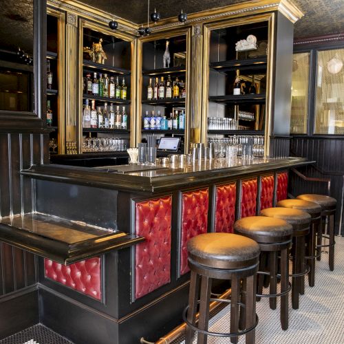 A stylish bar with red padded front panels, bar stools, shelves stocked with bottles, and a luxurious ceiling design in a well-lit space.