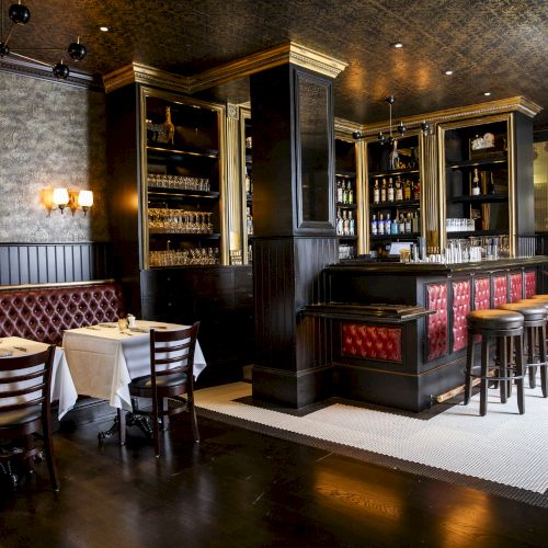 A stylish, dimly lit bar and restaurant features red cushioned seating, dark wooden decor, and a well-stocked bar with high stools and tables.