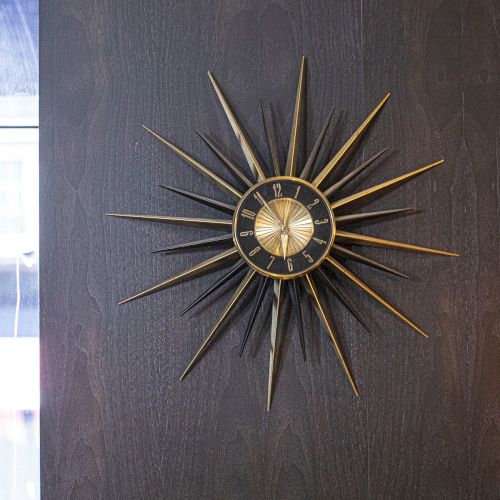 The image shows a sunburst wall clock with a black face and gold accents, mounted on a dark wall beside a window with a cityscape view.