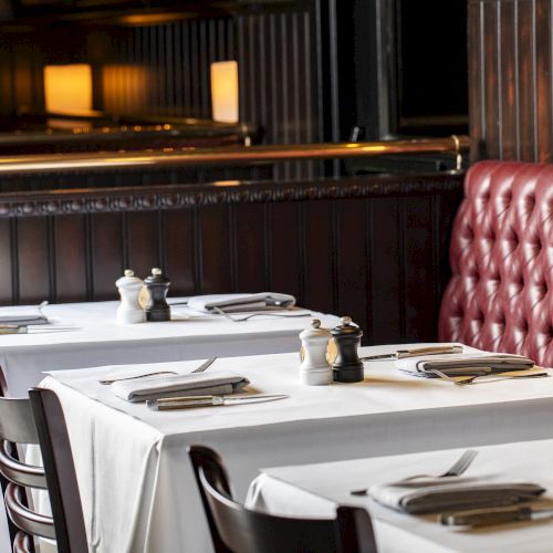 A cozy restaurant with white tablecloth-covered tables, red cushioned seating, neatly arranged cutlery, and salt and pepper shakers.