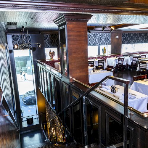 A wooden staircase leads to a well-lit dining area with white tablecloths, chairs, and window views.