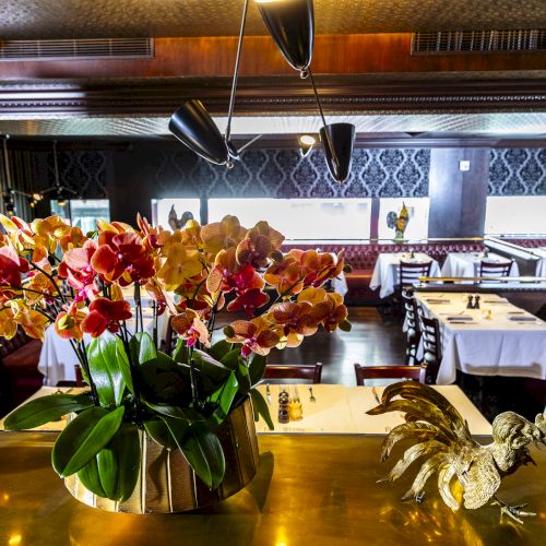 The image shows a restaurant interior with a floral centerpiece on a table alongside ornamental roosters, and tables set with white tablecloths.