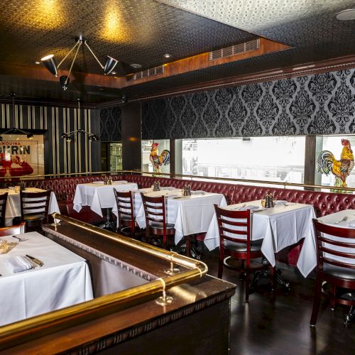 A restaurant with white tablecloths, wooden chairs, and a red upholstered banquette. Decor includes rooster paintings and patterned wallpaper.