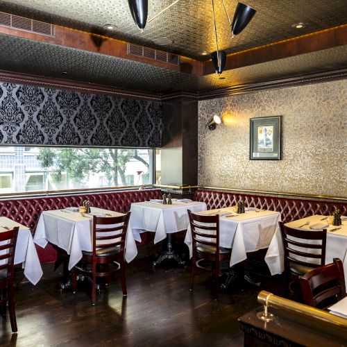 The image shows an elegant restaurant interior with neatly set tables, wooden chairs, patterned wallpaper, and framed art on the walls.