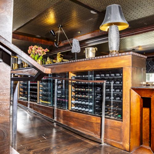 The image shows a stylish interior of a wine bar or restaurant with wine racks, wooden furnishings, a table lamp, and floral arrangements on the counter.