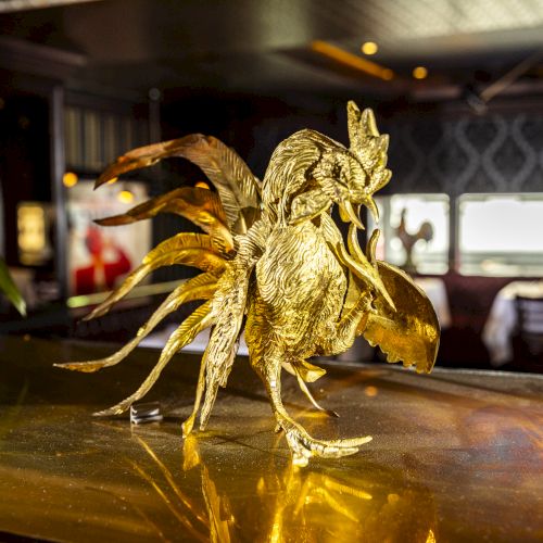 A golden rooster sculpture is displayed on a reflective surface in a dimly lit, elegant room with tables set in the background.