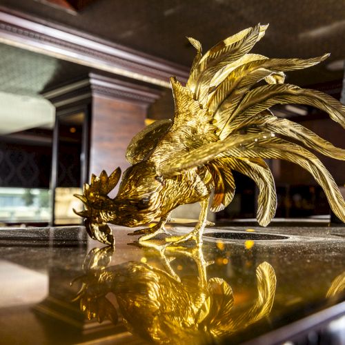 The image shows a golden rooster figurine placed on a reflective surface in an elegant setting, with ambient lighting and decor in the background.