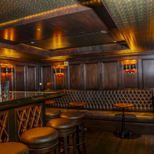 The image shows a cozy, dimly lit bar with wooden paneling, bar stools, a tufted leather bench, and small round tables.