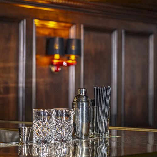 The image shows a bar counter with a cocktail shaker, stirring sticks, and a glass full of ice cubes in front of a wooden paneled wall with sconce lights.