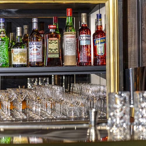 A bar setup with various liquor bottles on shelves, a row of wine glasses, and some cocktail equipment on the countertop, ending the sentence.