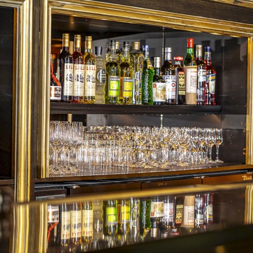 The image shows a bar with bottles of liquor and wine glasses neatly arranged on shelves, framed by a wooden cabinet with gold trim, ending the sentence.