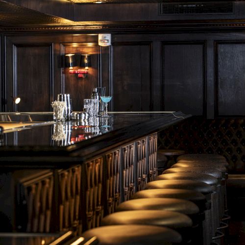 The image shows a dimly lit bar with multiple stools lined up along the counter, featuring glasses and bottles on the bar top.