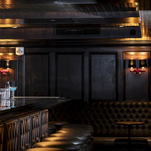 A dimly lit bar with cushioned seating, a row of stools by the counter, soft lighting fixtures, and various glassware on the bar top.