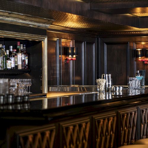 The image shows a dimly lit, elegant bar with a fully stocked liquor shelf, cushioned bar stools, and a polished counter adorned with glassware and shakers.