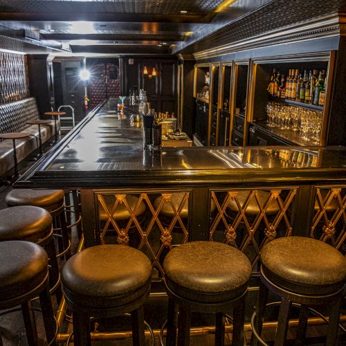 An empty bar with a polished counter, high stools, and shelves of alcohol bottles. The room has dark wooden decor and ambient lighting.