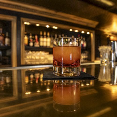 A glass of an orange-colored cocktail sitting on a napkin atop a glossy bar counter in a dimly lit bar with bottles and glasses in the background.