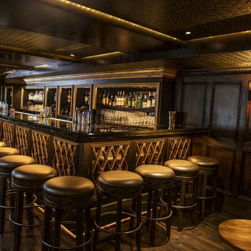 The image shows an empty, dimly lit bar with high stools placed around a long wooden counter, showcasing a variety of bottles and glassware.