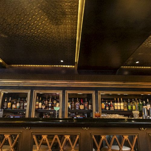 A sleek bar setup with a variety of liquor bottles on shelves, illuminated under a modern, gold-accented ceiling with dark tones, spotted lighting ending the sentence.