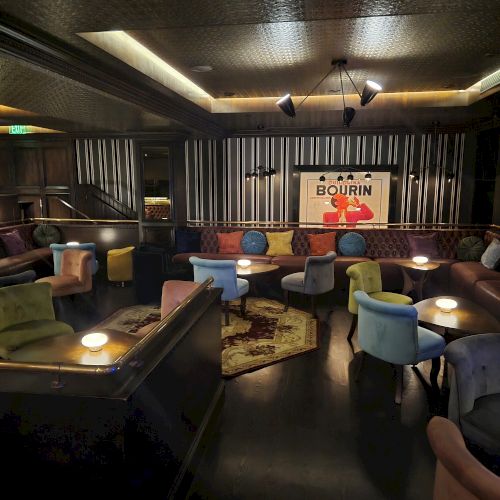 A cozy bar lounge with tufted booths, colorful chairs and small tables, plus a &ldquo;BOURRIN&rdquo; wall sign and spotlights overhead.