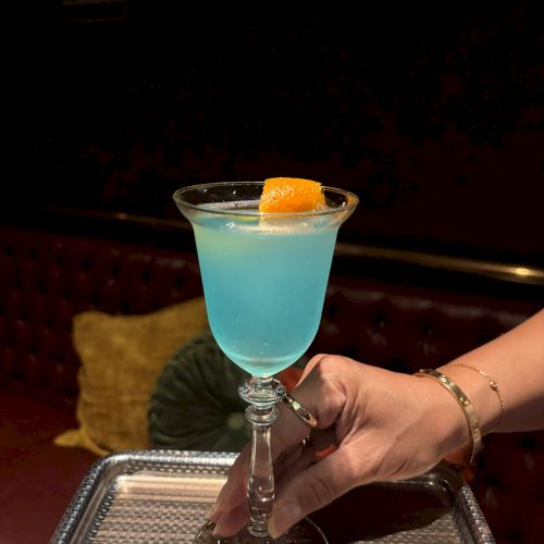 A hand holds a turquoise cocktail in a stemmed glass, topped with an orange cube garnish.