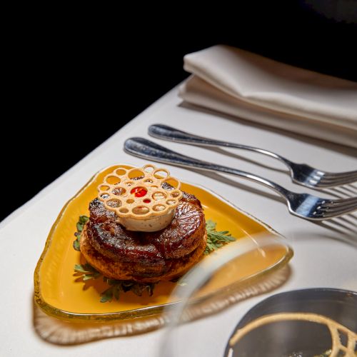 A plated steak with a decorative onion ring on an orange dish, with silver forks and a bread roll in the background.